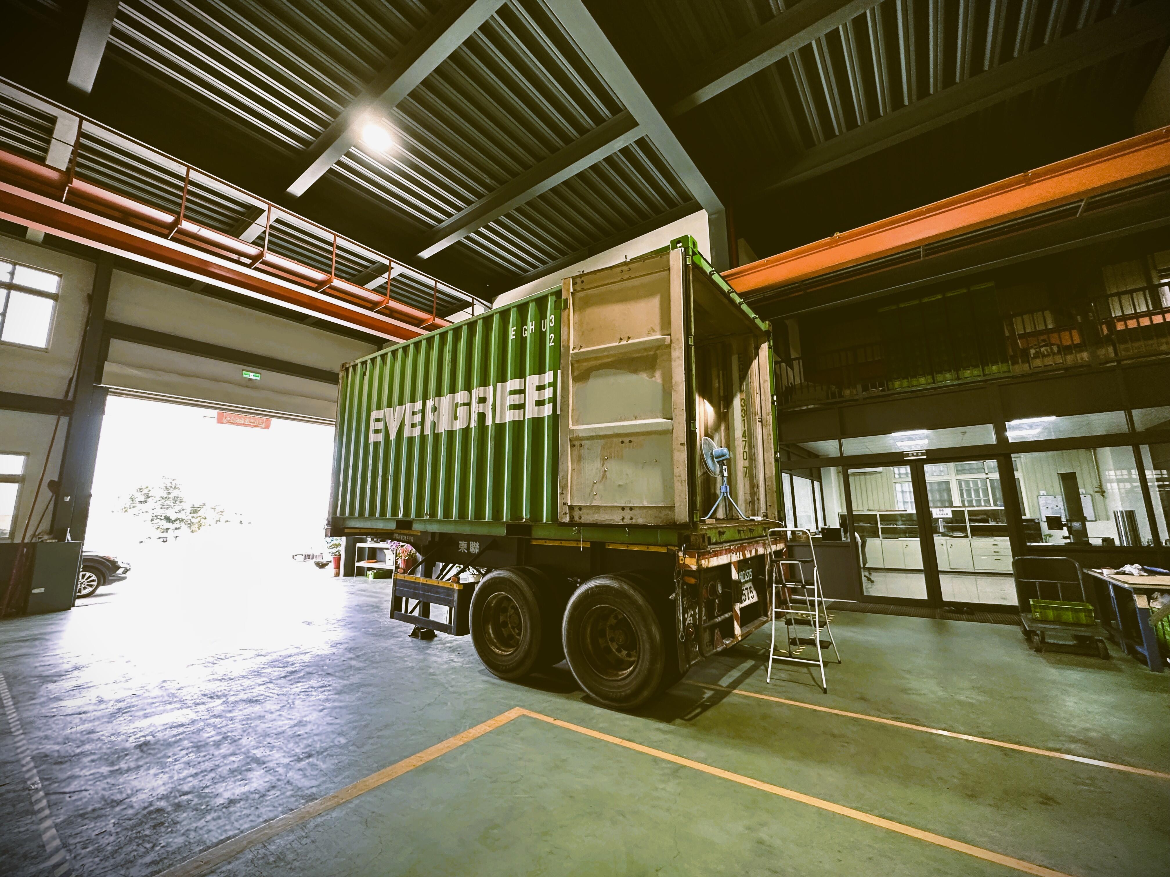 A large, green Evergreen shipping container on a truck trailer parked inside a warehouse or factory space.